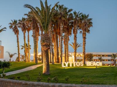 Palmen gesäumter Weg mit beleuchtetem "LYTTOS BEACH"-Schild bei blauem Abendhimmel.