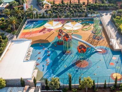 Bunter Spielplatz mit Rutschen und Klettergerüsten neben einem Hotel und viel Grün rundherum.
