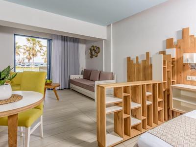 Jasno urządzony pokój hotelowy z częścią wypoczynkową, łóżkiem i widokiem na palmy przez okno.