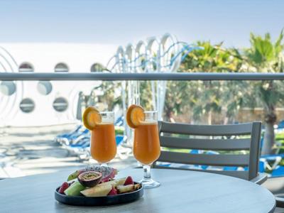 Balkon mit Tisch, Obstteller und zwei Cocktails mit Aussicht auf Pool und Palmen.