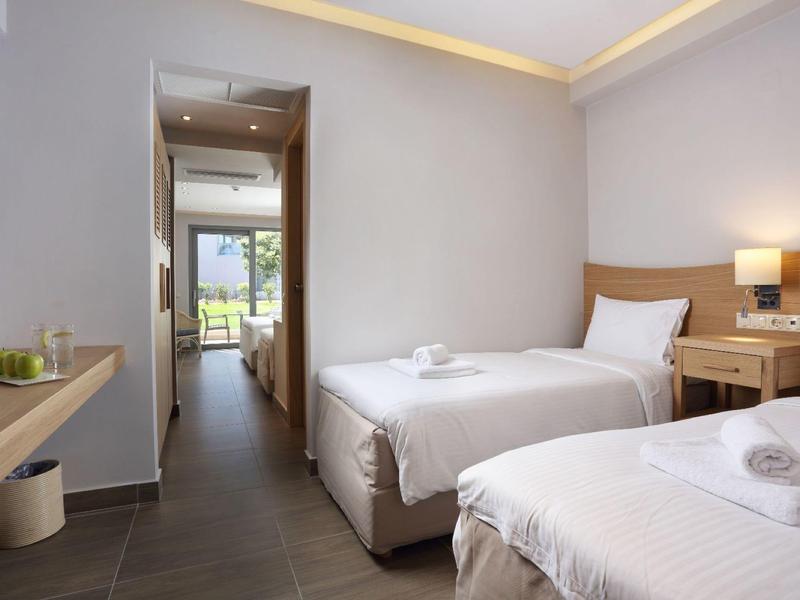Clean, minimalist twin room with twin beds, bedside table, lamp, and view to a balcony.