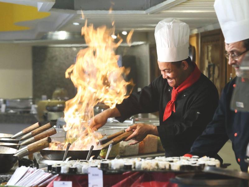 Chef cooking with a large flame in a restaurant kitchen, wearing white hats and black uniforms.