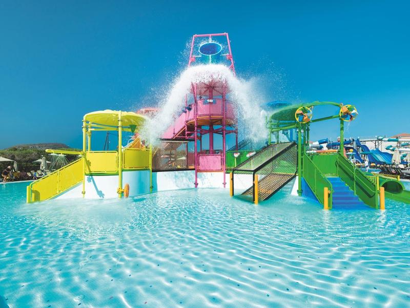 Water park with slides and splash bucket in a large pool under clear blue sky.