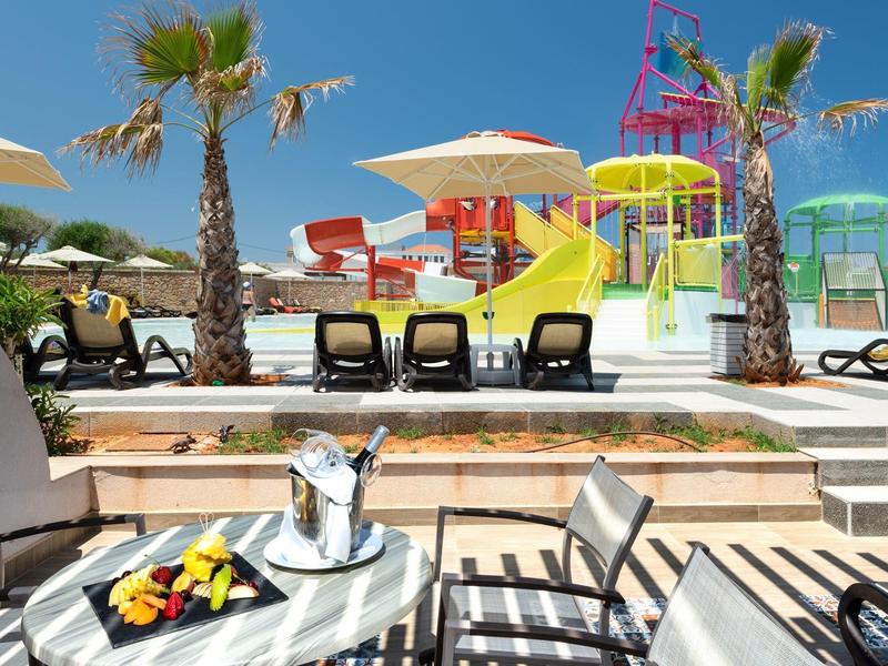 Hotel pool area with sun loungers, colorful water slides, palm trees, and a table set with fruit and drinks.