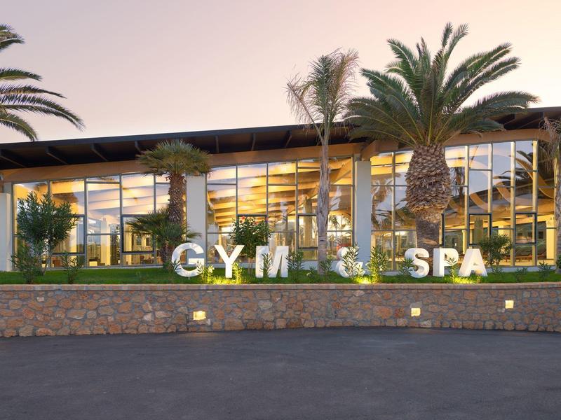 Modern gym and spa with glass walls, palm trees, and illuminated signs at dusk.