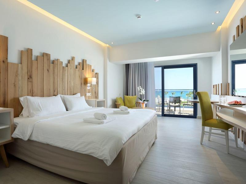 Bright hotel room with a large bed, wooden headboard, desk, and balcony overlooking the sea.