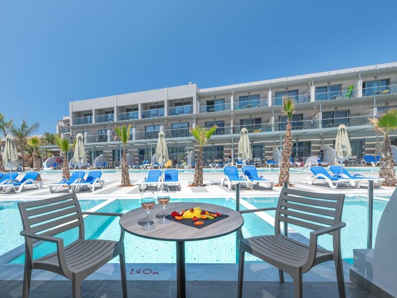 Resort pool with sun loungers, umbrellas, and a table with fruit near a modern hotel under clear sky.