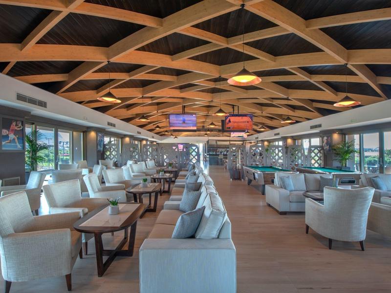 Spacious hotel lounge with modern armchairs, wooden tables, large windows, and decorative ceiling.