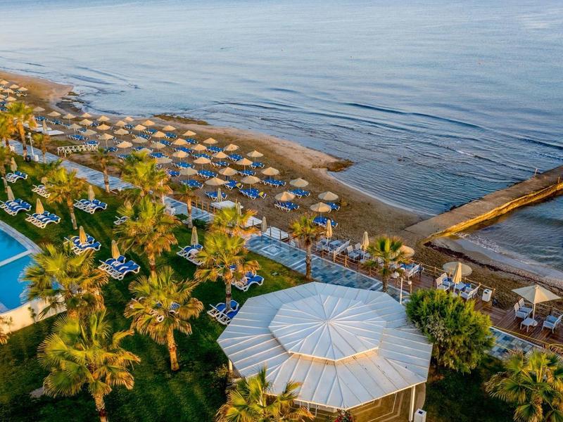 Aerial view of a beach resort with umbrellas, pool, palm trees, and calm sea water.