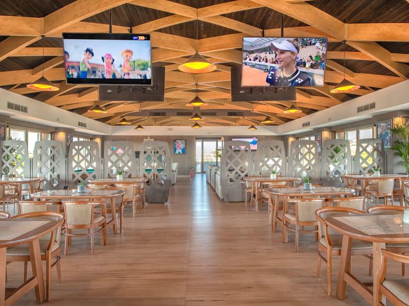 Spacious hotel restaurant with wooden furniture, geometric ceiling, and TV screens.