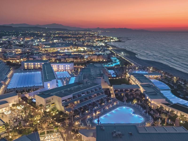 View of a lit luxury hotel with pools and sea at sunset.
