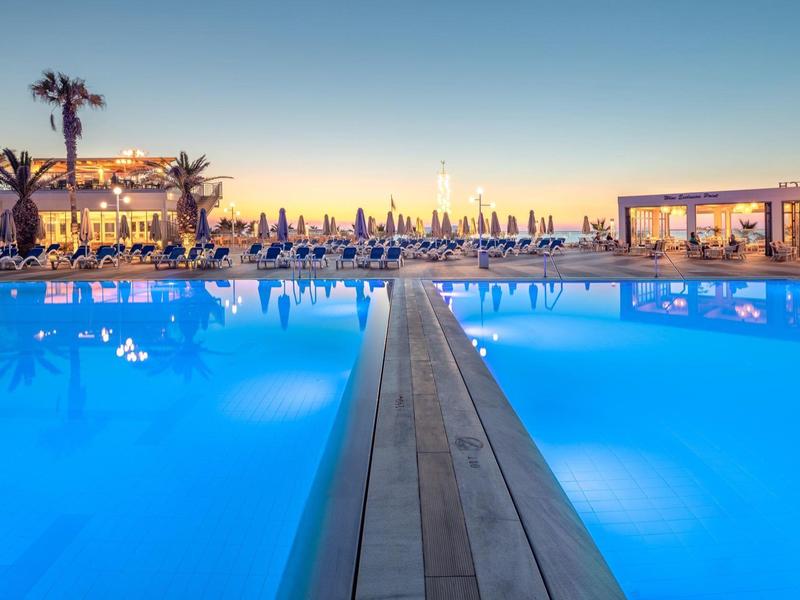 Sunset view of a large poolside area with umbrellas and palm trees at a resort.