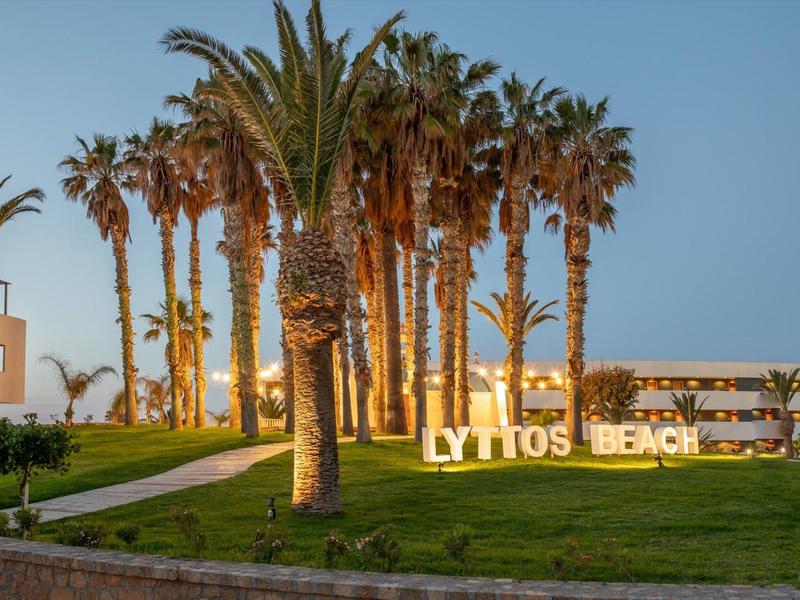 Palm trees line a path leading to a beach resort with 'LYTTOS BEACH' sign lit up at dusk.