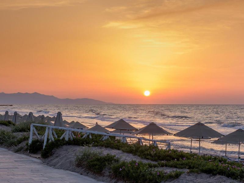 Sunset over a beach with umbrellas and calm sea, mountains in the background.