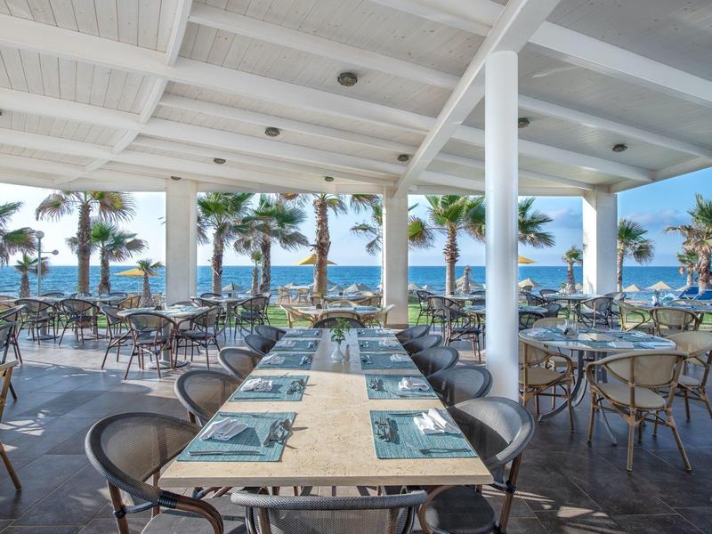 Open-air restaurant with tables and chairs overlooking palm trees and the ocean under a white roof.