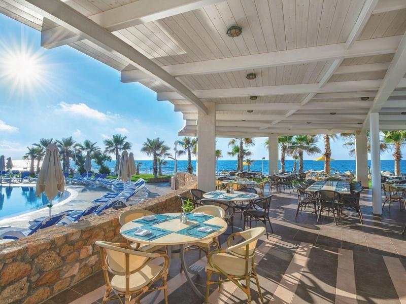 Terrace with tables and chairs beside a pool with palm trees and ocean under sunny weather