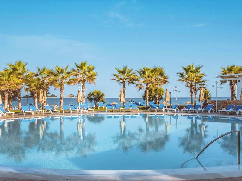 Large pool with lounge chairs and palm trees under a blue sky by the sea.