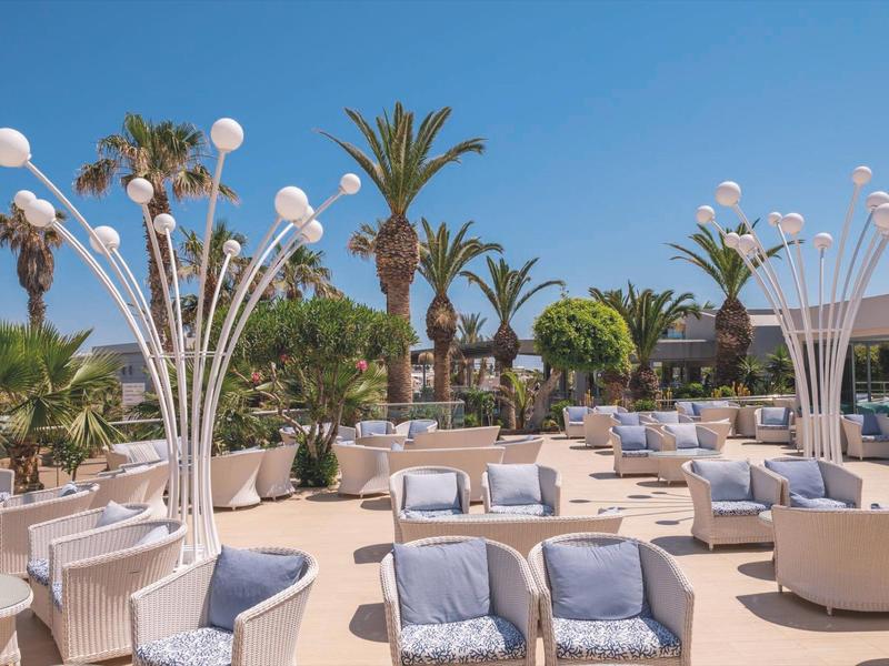Bright modern outdoor lounge with armchairs, palm trees, and decorative white lamps under clear sky.