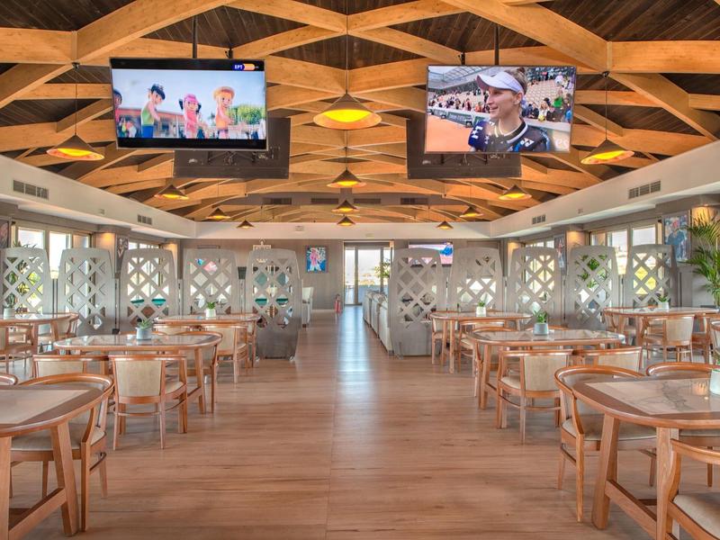 Large, bright dining room with wooden tables, chairs, and two ceiling-mounted screens.