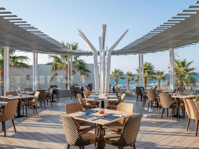 Open-air restaurant with sea view, rows of tables, palm trees, and modern pergola roof.