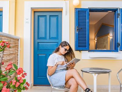Une femme est assise sur une chaise devant une maison avec une porte bleue et une fenêtre ouverte en lisant un livre.