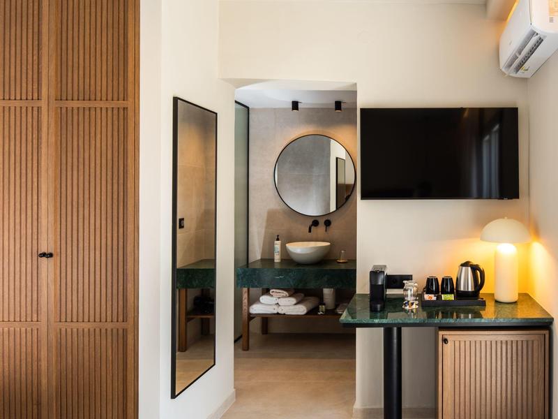 Area di camera d'hotel moderna con armadio in legno, grande specchio, bagno con specchio rotondo e TV sopra un mobile.