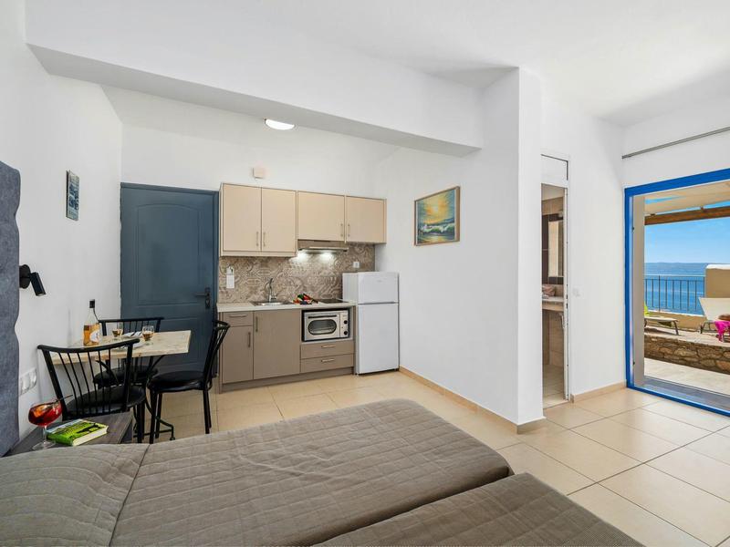 Jasna kuchnia w pokoju hotelowym z lodówką, stołem, łóżkiem i widokiem na taras z morzem.