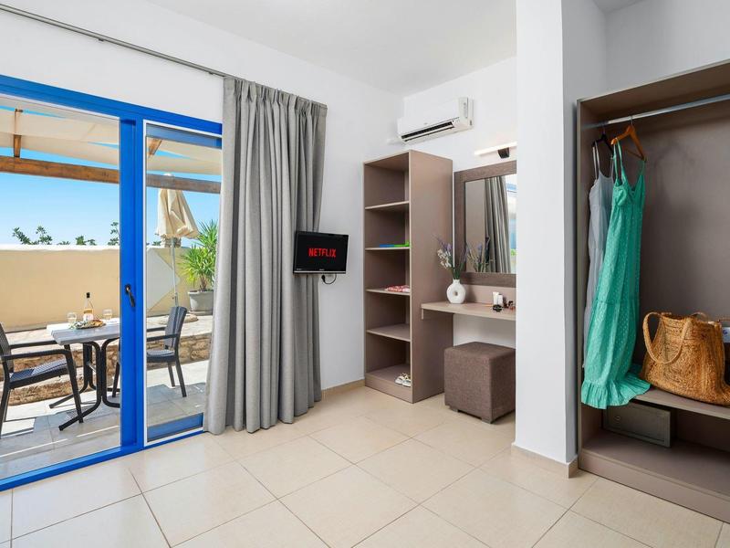 Jasny pokój hotelowy z garderobą i wyjściem na taras z krzesłami i parasolem.