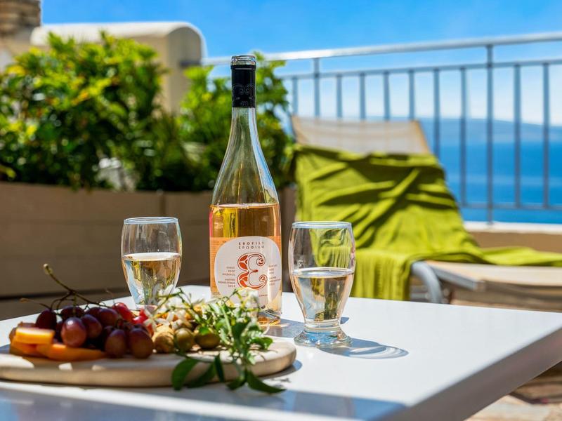 Butelka wina i dwie szklanki na stole z widokiem na morze i zielonymi poduszkami w tle.