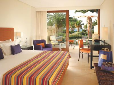 Habitación de hotel luminosa con cama grande, sillones y vista a una terraza con palmeras.