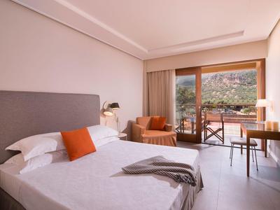 Jasny pokój hotelowy z podwójnym łóżkiem, pomarańczowym fotelem, biurkiem i balkonem z widokiem na zieleń.