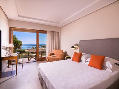 Habitación de hotel luminosa con balcón y vista al mar, equipada con una cama grande y muebles modernos.