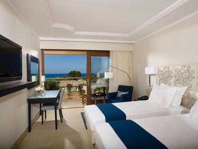 Habitación de hotel luminosa con dos camas, TV, escritorio y balcón con vista al mar.