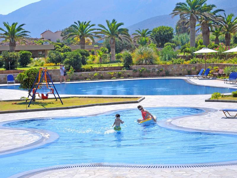 Basen na zewnątrz z wyspą i bawiącymi się dziećmi, otoczony palmami i parasolami.