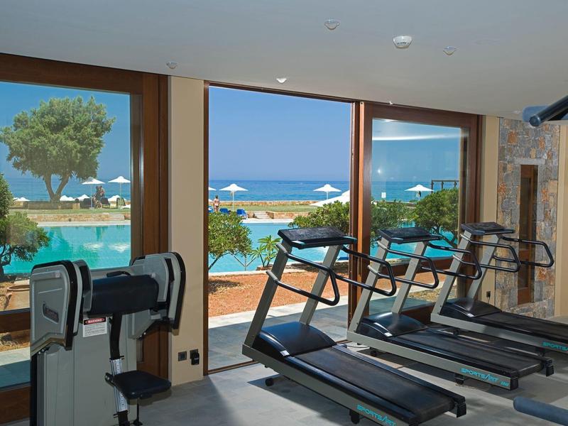 Siłownia z bieżniami z widokiem na basen i morze z parasolami i drzewami za dużymi oknami.