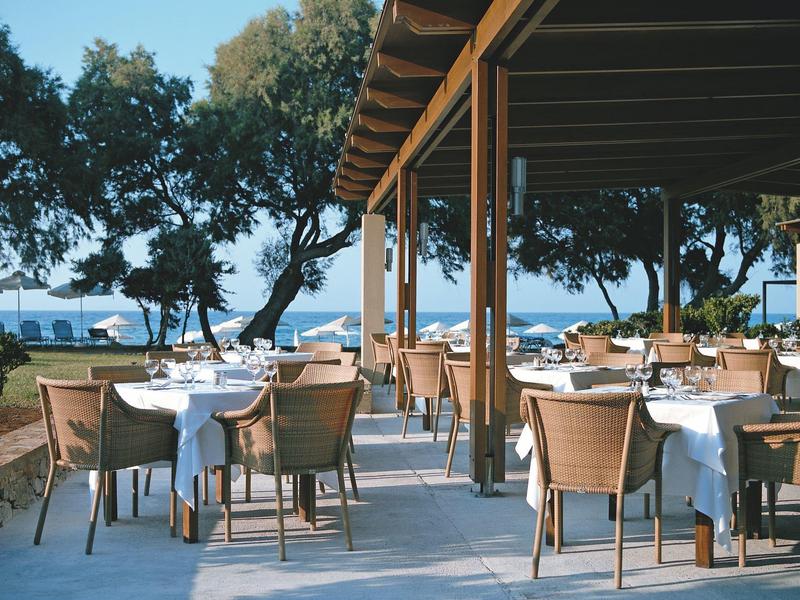Zewnętrzny taras jadalny pod zadaszeniem z stołami i krzesłami obok drzew i widokiem na morze.