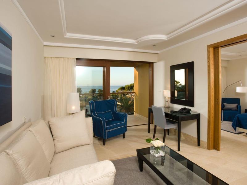 Nowoczesny pokój hotelowy z beżową sofą, niebieskim fotelem, biurkiem i szklanymi drzwiami na balkon.