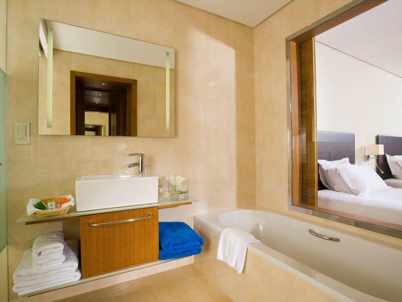 Nowoczesna łazienka z beżowymi ścianami, wanną, umywalką, ręcznikami i oknem na sypialnię.