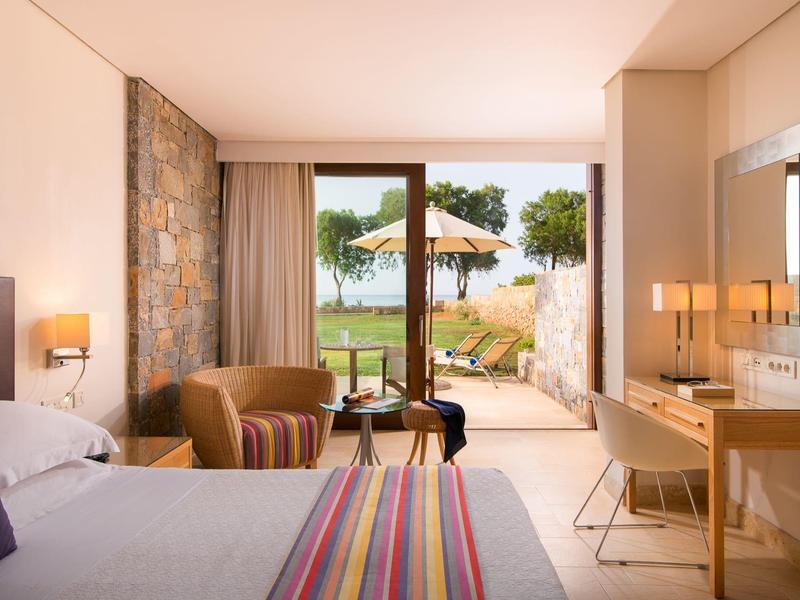 Pokój hotelowy z łóżkiem, fotelem, biurkiem i drzwiami przeszklonymi otwierającymi się na ogród z parasolami.