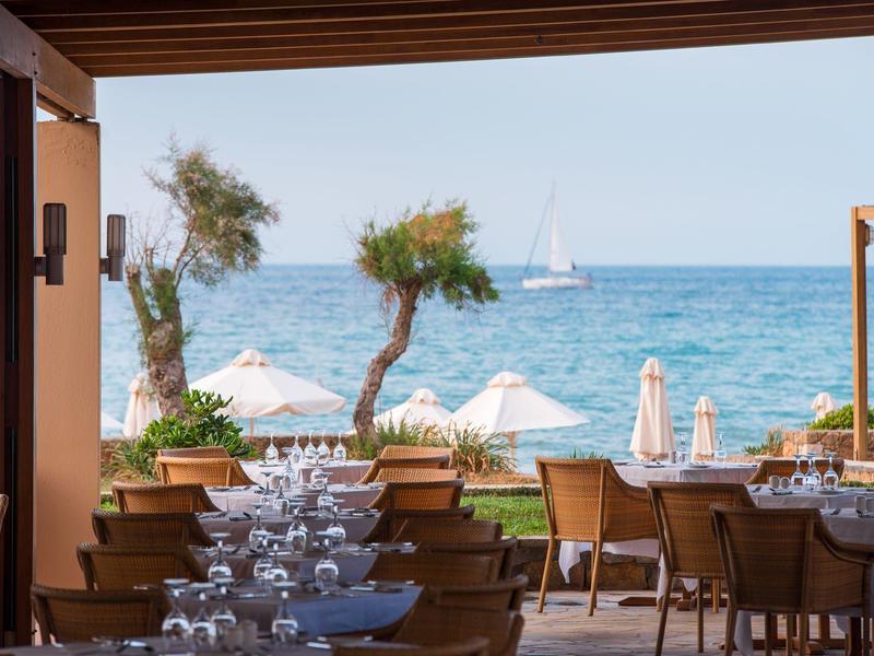 Restauracja nad morzem z nakrytymi stolikami, widokiem na morze, drzewami i żaglówką na horyzoncie.