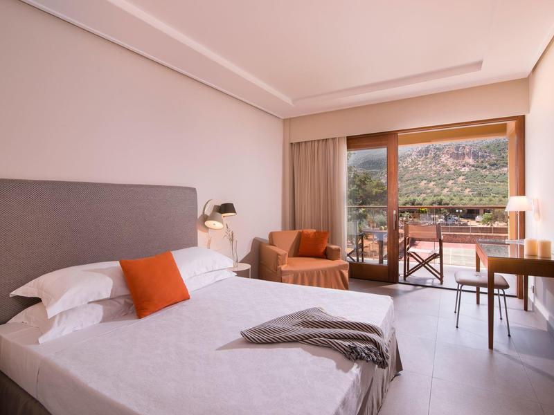 Jasny pokój hotelowy z podwójnym łóżkiem, pomarańczowym fotelem, biurkiem i balkonem z widokiem na zieleń.