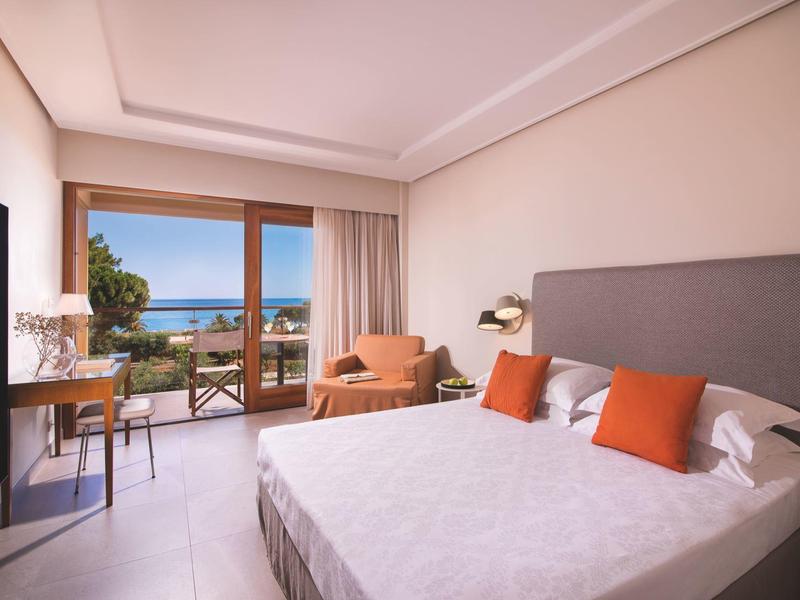 Jasny pokój hotelowy z dużym łóżkiem, biurkiem i balkonem z widokiem na morze.