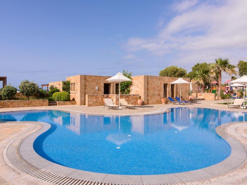 Słoneczny basen na zewnątrz hotelu z leżakami i parasolami pod jasnym niebem.