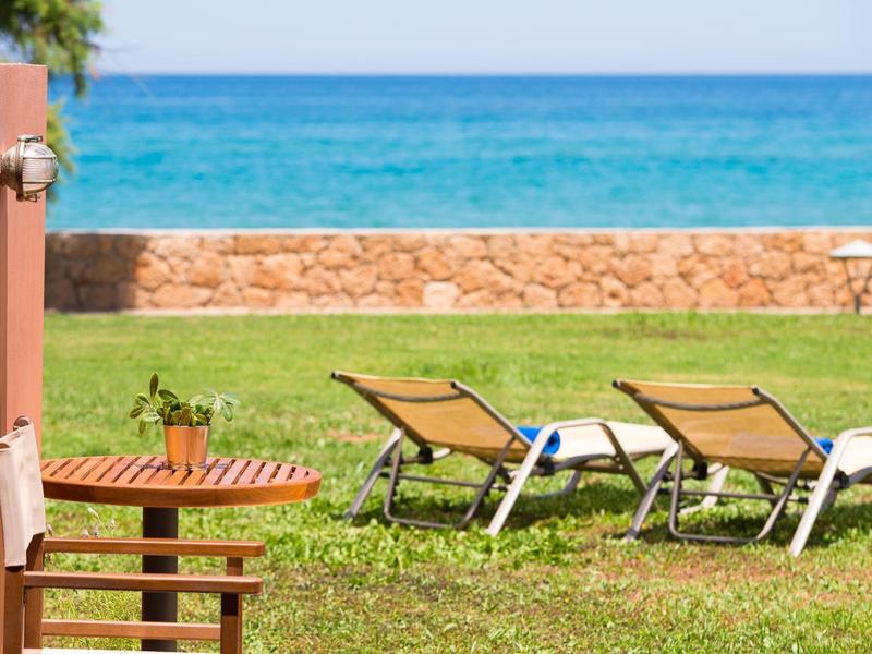 Dwa leżaki na zielonej trawie z widokiem na niebieskie morze, kamienny mur i czyste niebo.