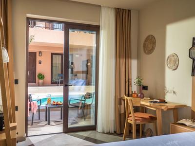 Helles Zimmer mit Holzmöbeln, Schreibtisch, Stuhl, Bett und Blick auf Terrasse mit Pool.