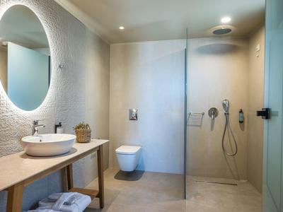 Modernes Badezimmer mit runder beleuchteter Spiegel, Holzkonsole, hängendem WC und Glas-Duschkabine.