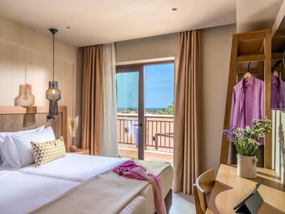 Camera d'hotel luminosa con due letti, balcone con vista mare e scrivania con specchio e fiori.