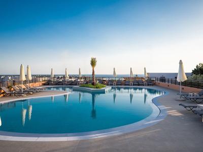 Runder Pool mit klar blauem Wasser, Sonnenschirmen und Stühlen, dahinter Meer und blauem Himmel.