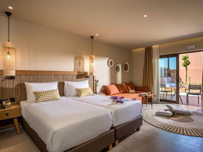 Bright hotel room with two beds, lounge area, and balcony overlooking palm trees.