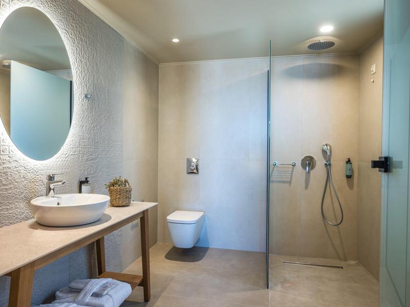 Modernes Badezimmer mit runder beleuchteter Spiegel, Holzkonsole, hängendem WC und Glas-Duschkabine.